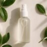 A hand holding a clear facial mist spray bottle against a soft neutral background